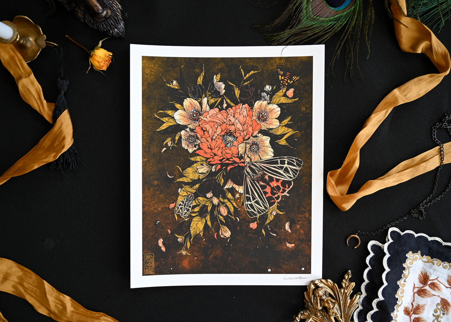 A photo of a 9x12 print on a black table. There are ribbons, candles, and hankerchiefs on the table. The print is Bloom Lepis. It shows three moths perching on over blooming flowers.