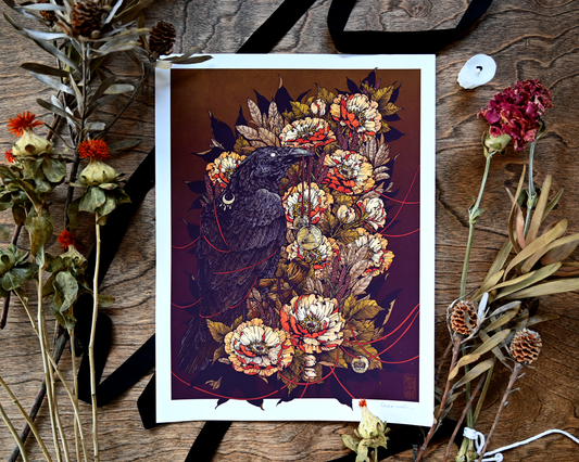 A photograph of a giclee print sitting on a wood table. Under the print is a black silk ribbon. Around it is dried flowers. The print shows a black crow tangled in a red sting. The crow is perching on a bouquette of flowers.