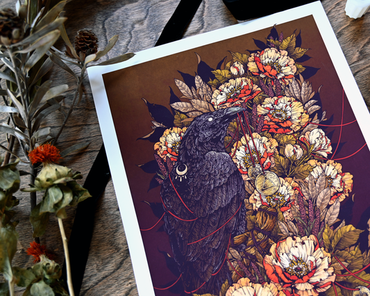 An up close photo of the print. It is on a diagonal from the left to the right. It shows the details of the top half of the print. You can see how each feather is rendered and what the coins in the raven's mouth looks like. As well as the flowers behind the crow. You can also see the dried flowers on the table. 