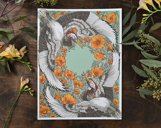A photograph of a giclee print sitting on a darkwood table. There are cut fresh flowers on the table around the print. The print depicts two cranes engaged in an elegant dance. Around them are orange california poppies.