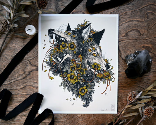 A photo of a giclee print sitting on a table. Around the print is black ribbon, an unlit candle, a crystal, and dried plants. The print shows two wolf heads attached to each other. The wolves have far too many eyes amd there is flowers bursting out of them. 