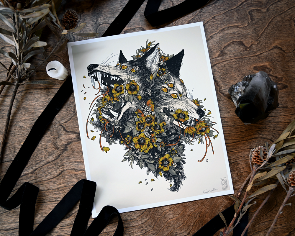 A photo of a giclee print on a dark wood table. You can see the print on the left to right diagonal. You can see the whole print of the conjoined wolf heads being taken over by flowers. There is ribbon, a candle, a crystal, and dired plants on the table.