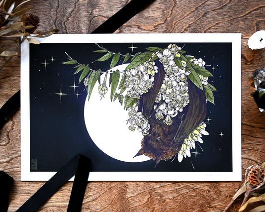A photo of a giclee print on a table. There is a black ribbon, an unlit candle, and dried plants. The print is of a flying fox bat tucked into durian flowers. Behind them is in front of a full moon.