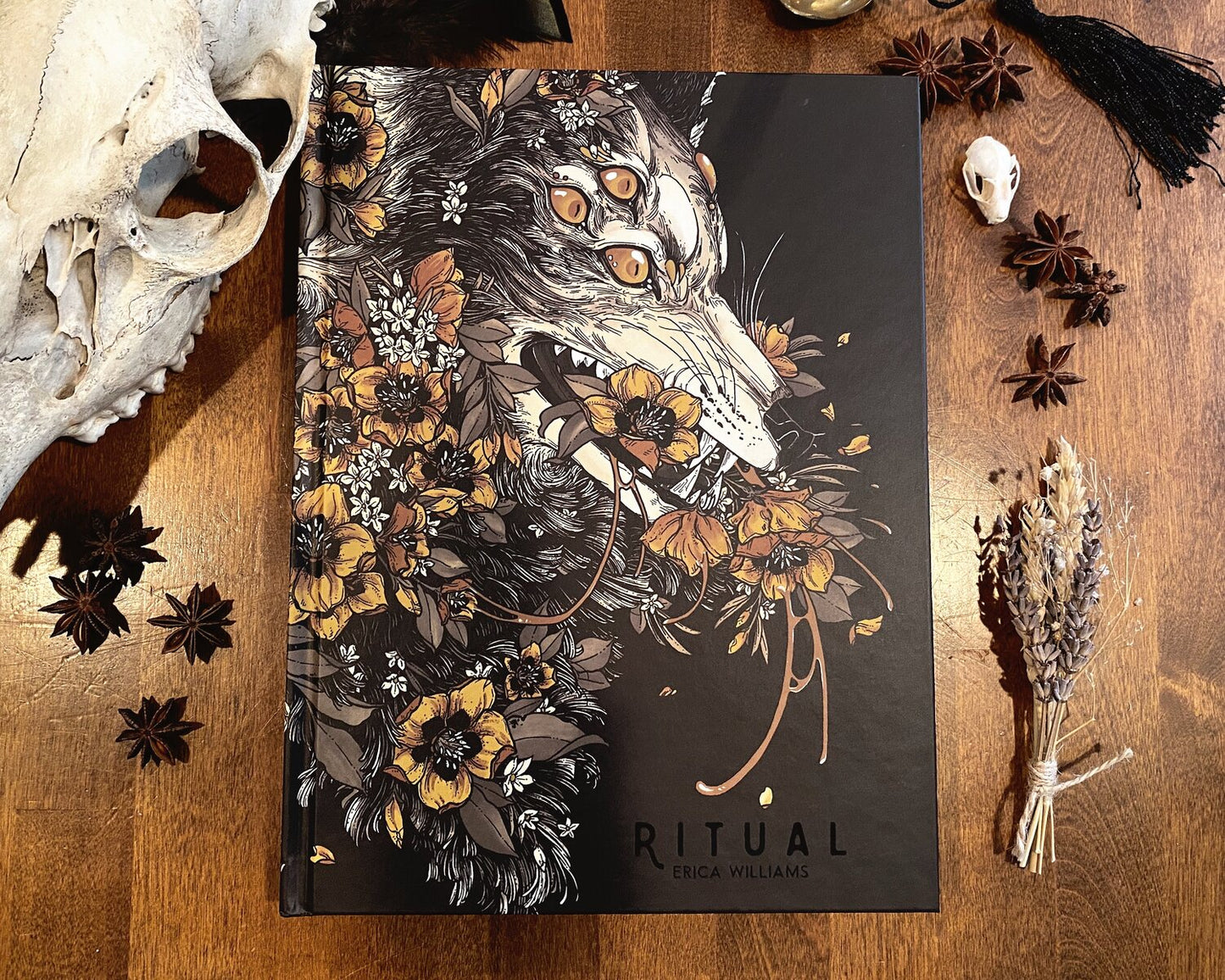 Photograph of the art book ritual. The cover shows a multieyed wolf overgrown with flowers. On the table are animal skulls, star anise, and a bundle of lavender.