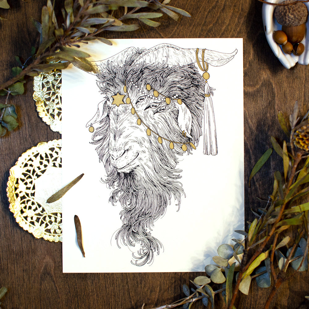 A photograph of a screen print on a table. Under the print is is gold paper doilies. Around the print is dried leaves, and acorns. The print shows a goat with 6 eyes, a star of david on his forhead, and chains hanging down over his forehead. There is a tassle hanging from his horn.