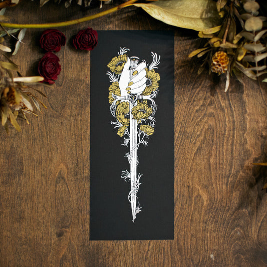 A photograph of a long and narrow screen print on a wood table. There are dried flowers above the print. The print shows a feminine hand with gold nails clutching a sword. Around the hand and stiletto are gold poppies.