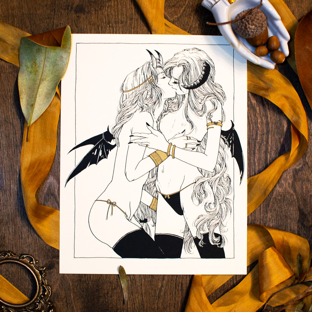 A photo of a screen print on a wood table. There is gold ribbon under the print. Also on the table is ceramic hands holding acorns, a leaf, and a small gold frame. The print is of two demon ladies kissing with gold acents.