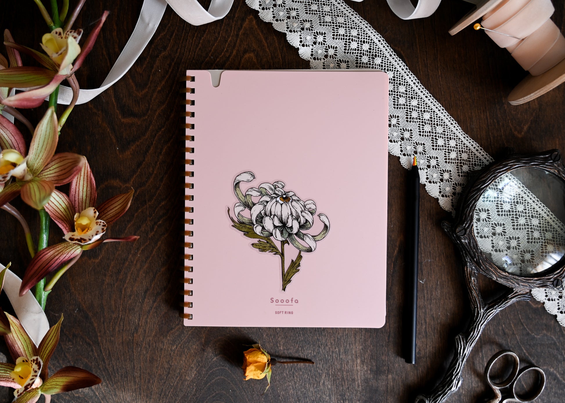 A photograph showing a pink notebook on the center of the table. There is lace, ribbon, faux flowers, a magnifying glass and a pair of scissors. The pink notebook has the chrysanthemum sitckers applied to the center of it.