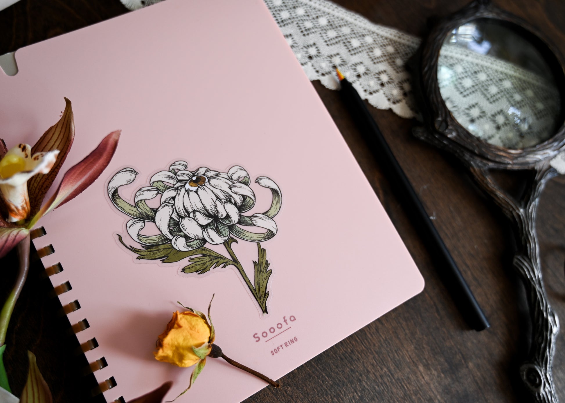 A photo showing the sticker applied to a pale pink notebook. The notebook is sitting on a darkwood table. There is a dried yellow rose sitting on the notebook. There is faux flowers, a marker, lace, and a magnifying glass. 