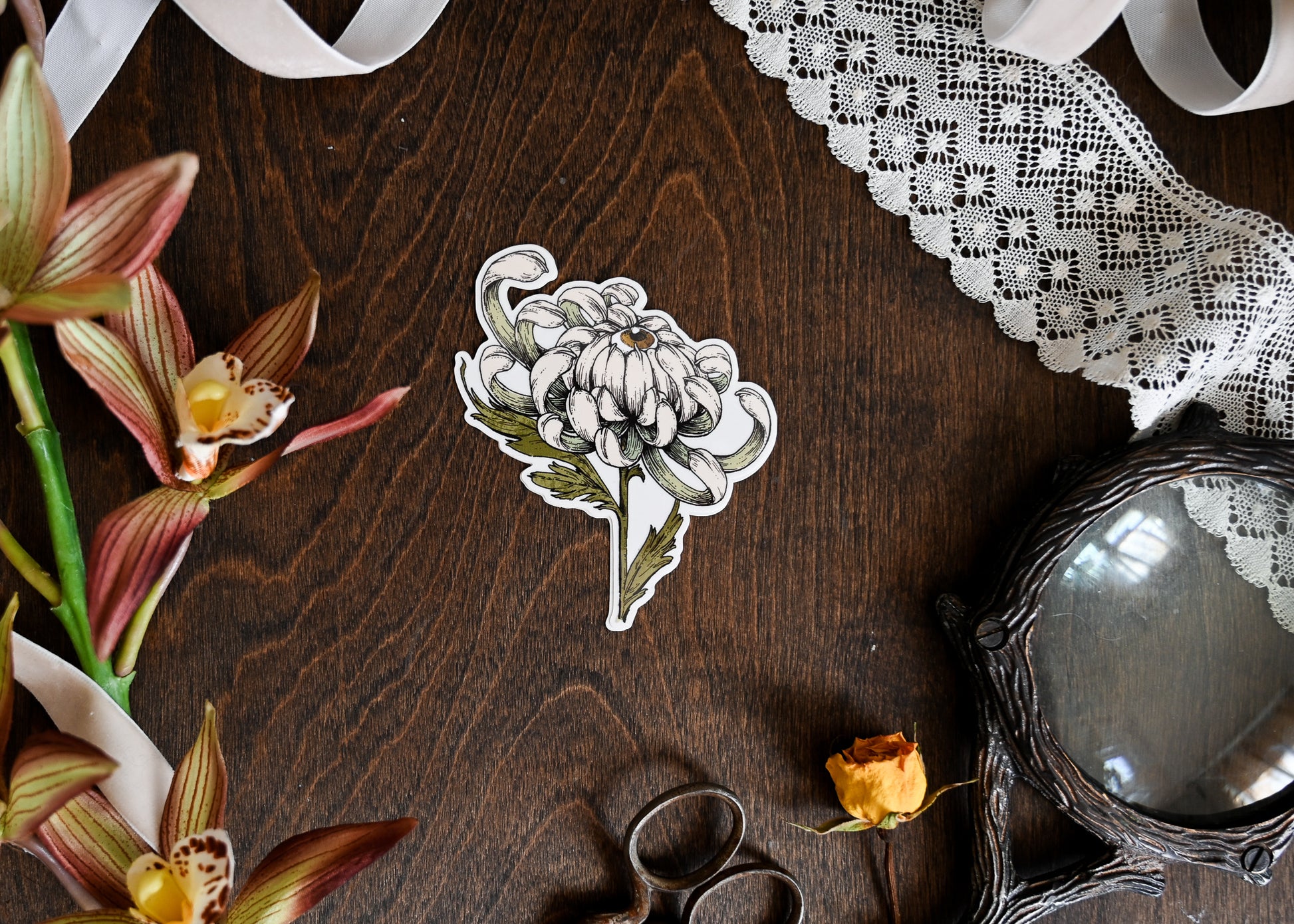 A photograph of a clear back sticker sitting in the center of a dark wood table. The sticker features an eyeball poking out of a chrysanthemum. There is lace, a magnifying glass, and faux flowers.