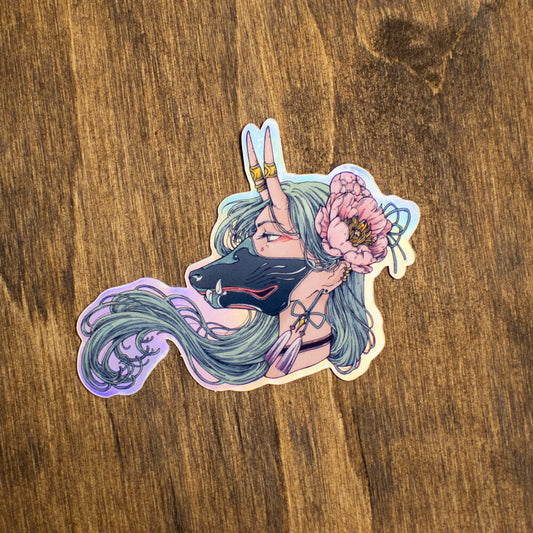 Photo of a weather proof holographic sticker on a wood table. The sticker shows a horned woman wearing a dog mask. There is a large flower in her hair. 