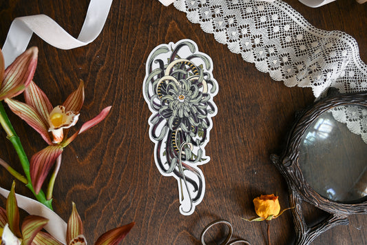 A photograph of a sticker on a table. Around the sticker is lace, ribbon, faux flower, sissors, and a magnifying glass. The sticker is of the so below art. A mutated chrysanthemum with an eye in the center.
