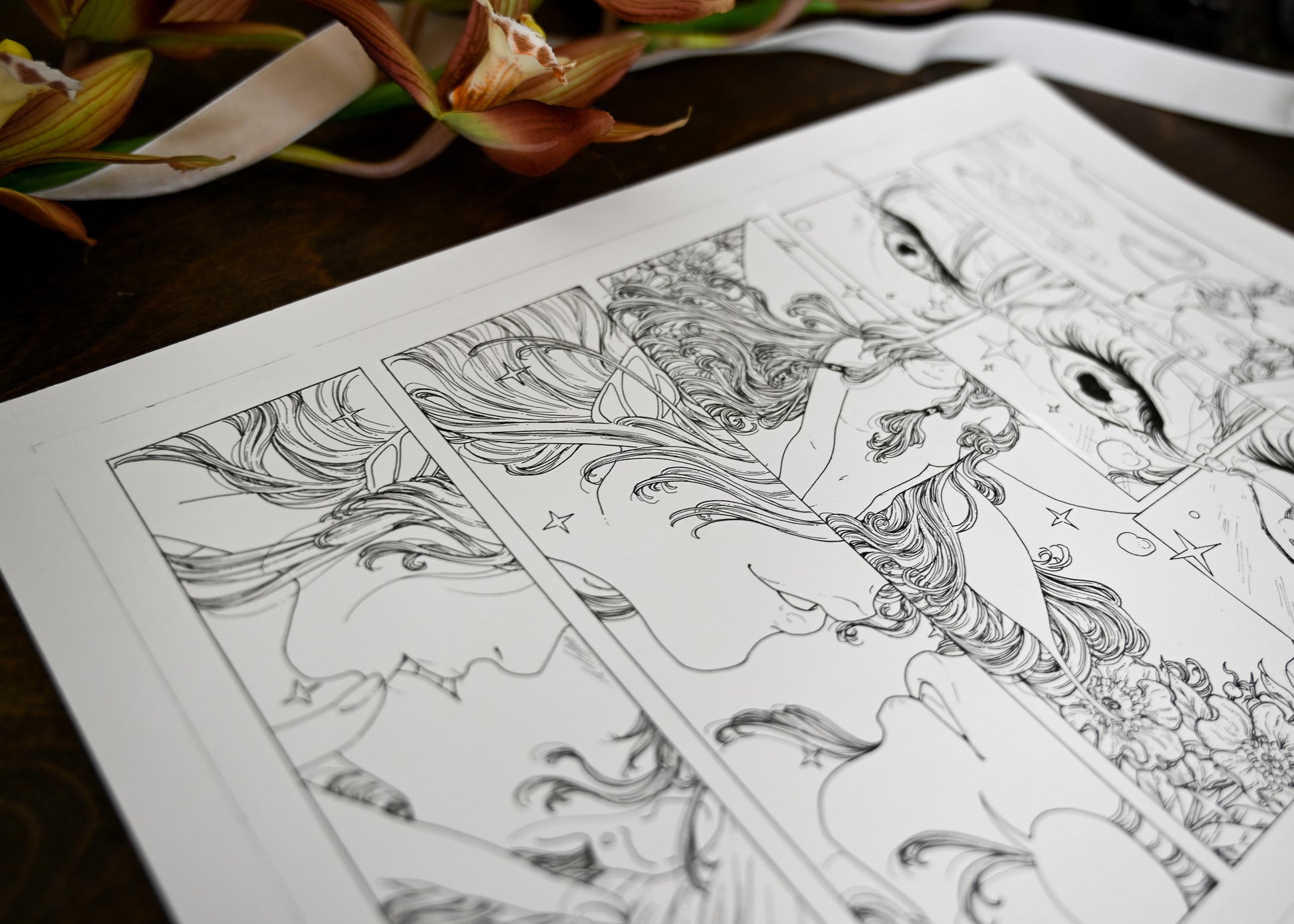 An up close photograph of a comic page. It shows the crisp and delicate ink lines. The drawing of the woman kissing the fairy is clear and the rest of the page is blurry. You can also see the fake orchids and ribbon on the left upper corner of photo. 