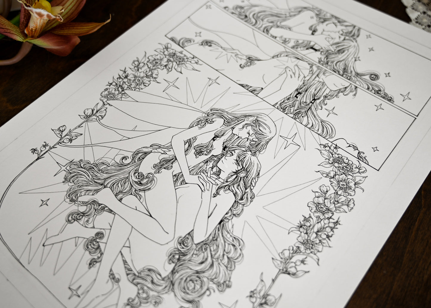 An up close of the comic page of the fairy and woman kissing and being intimate. You can clearly see the woman and fairy sitting together. The rest of the page is blurred. You can also see a fake orchid on the upper left corner. 